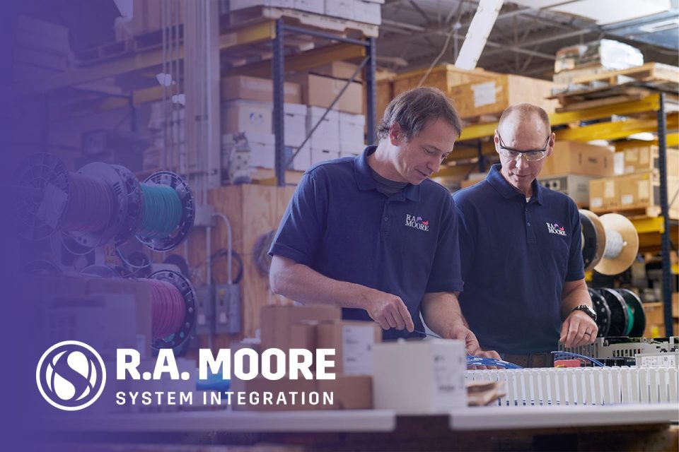 Two male employees of R.A. Moore dressed in purple polos with the R.A. Moore logo an the left breast work on a piece of system integration equipment with dark gradient overlay