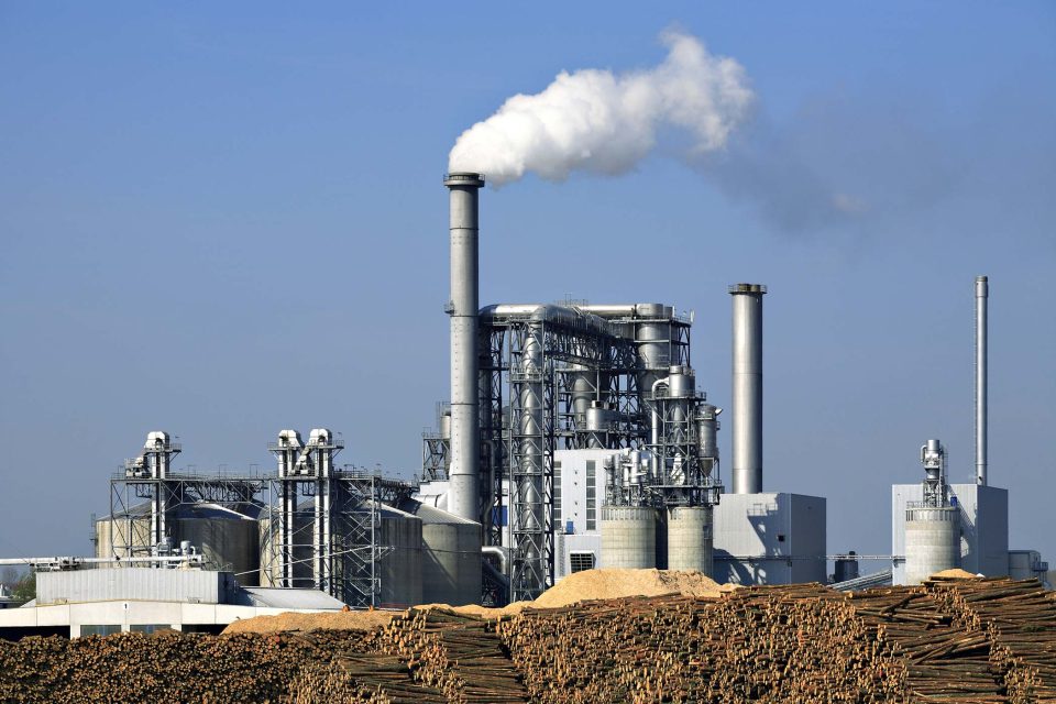 Paper manufacturing plant with long silver stacks issuing smoke and piles of lumber in the foreground.