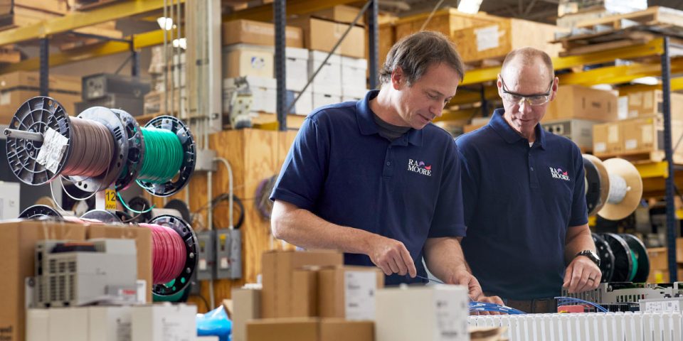 Two male employees of R.A. Moore dressed in purple polos with the R.A. Moore logo an the left breast work on a piece of system integration equipment